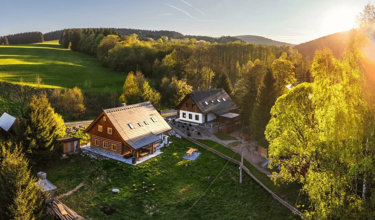 AI fotka stejné chaty v létě z dronu — zelená louka, ranní slunce. Sezónní varianta pro Airbnb listing, vytvořená AI z jediné originální fotky.