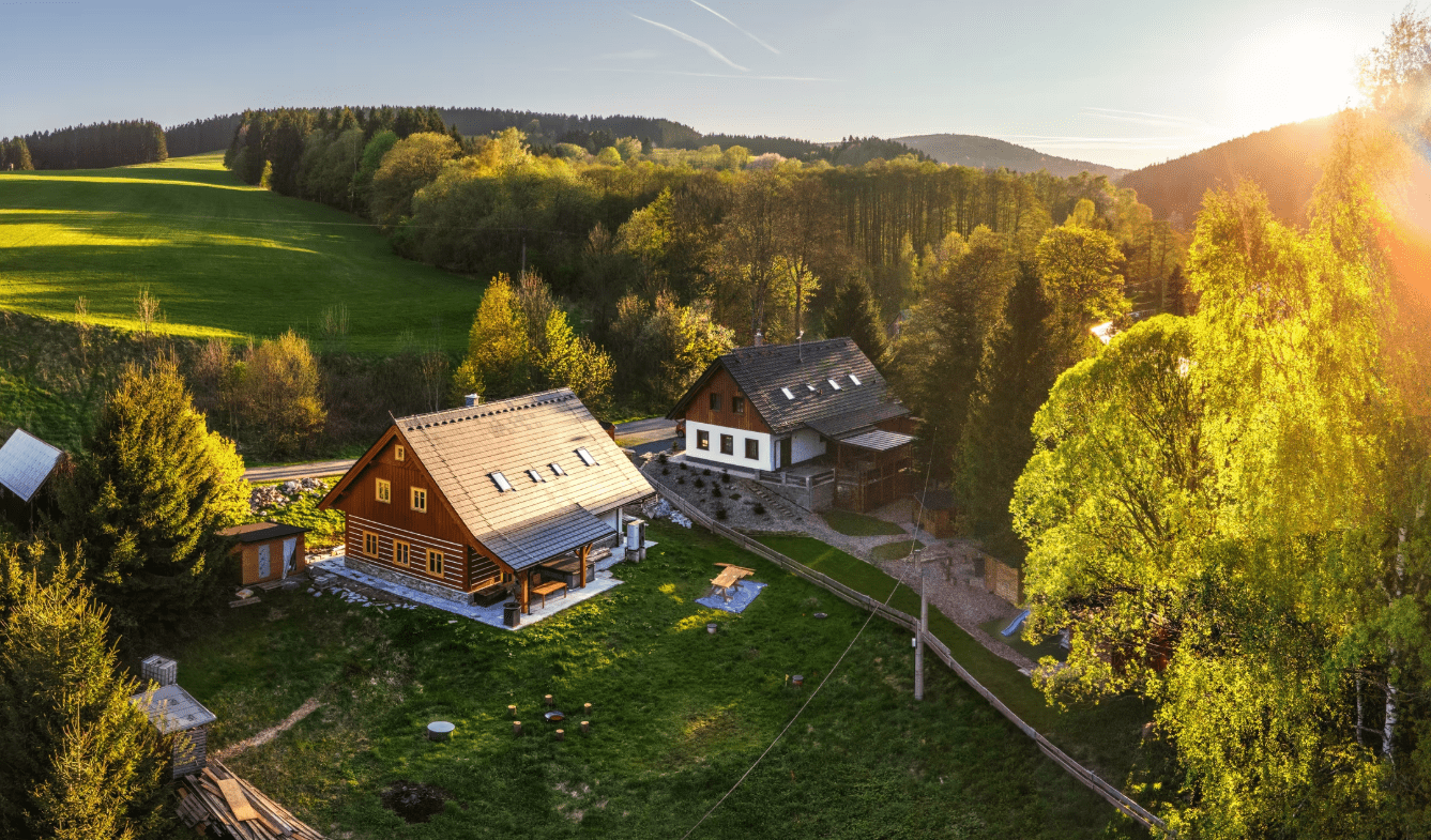 AI fotka stejné chaty v létě z dronu — zelená louka, ranní slunce. Sezónní varianta pro Airbnb listing, vytvořená AI z jediné originální fotky.